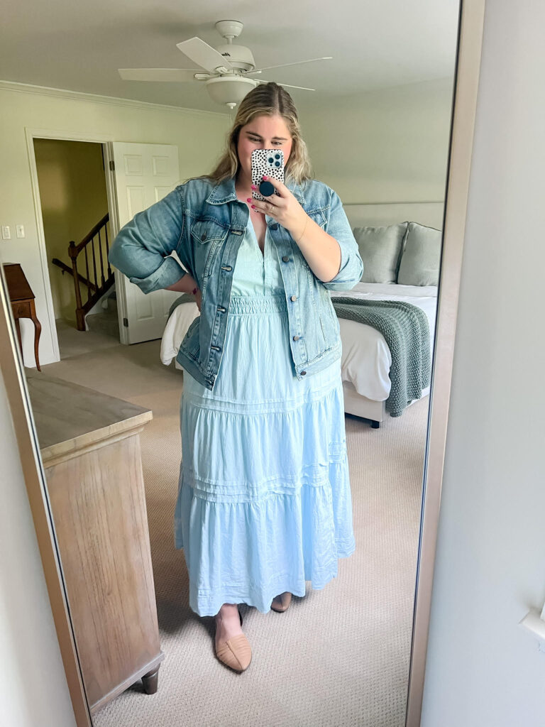 A Caucasian woman is taking a mirror selfie while wearing a light blue full length dress and light wash denim jacket. 