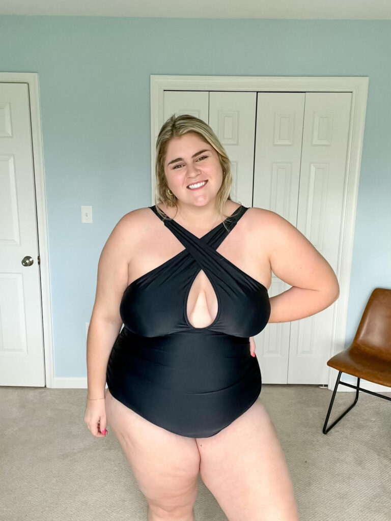 A happy plus size blonde woman wearing a plus size black one-piece bathing suit in her bedroom. 