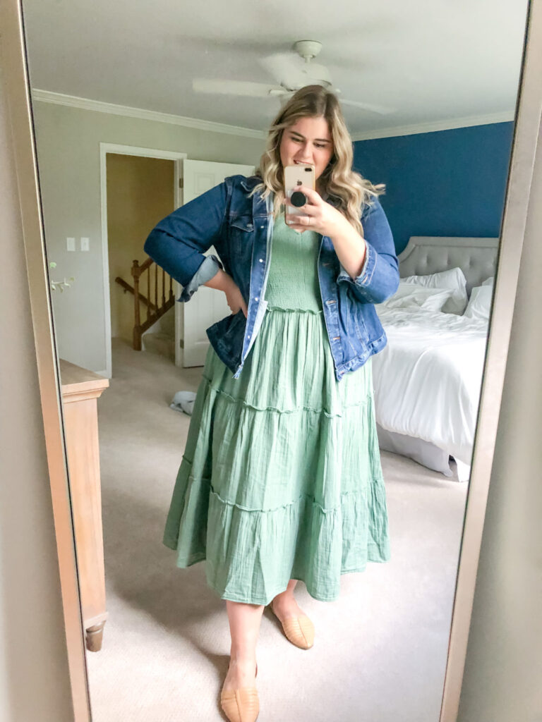 A plus size blonde woman is taking a mirror selfie and wearing a green tiered dress and denim jacket. 
