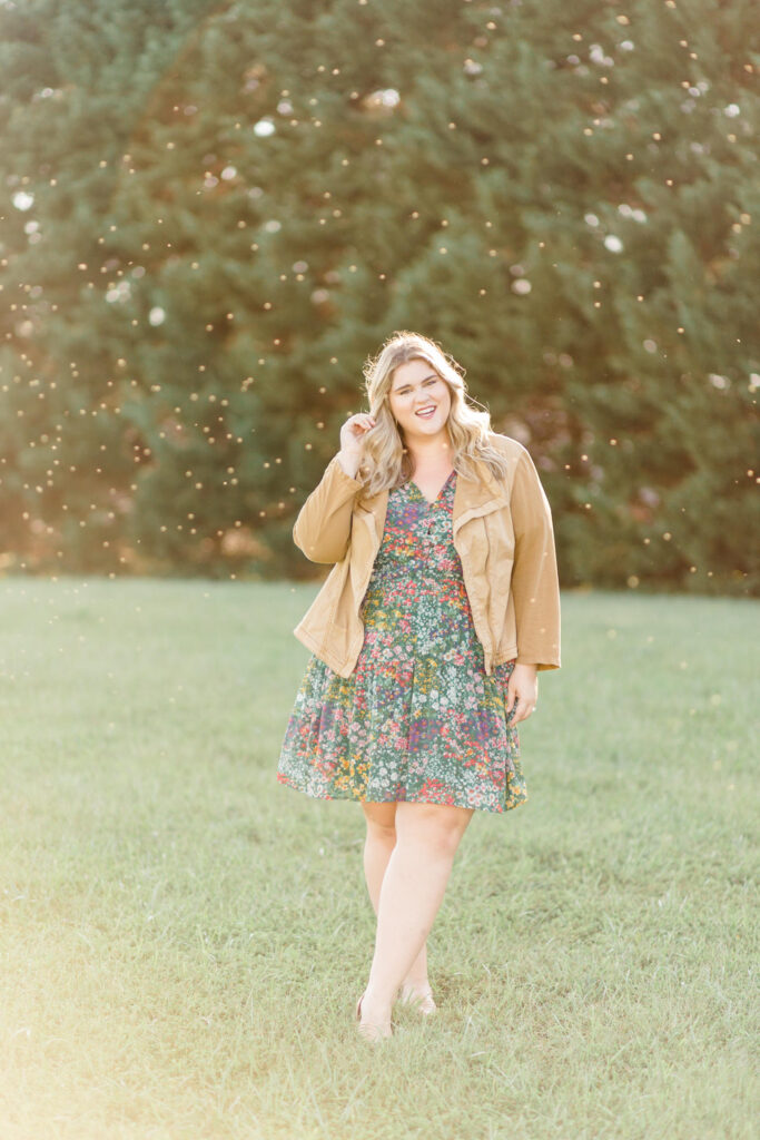 A plus size blonde woman walking outside in the grass wearing a floral dress and brown utility jacket. 