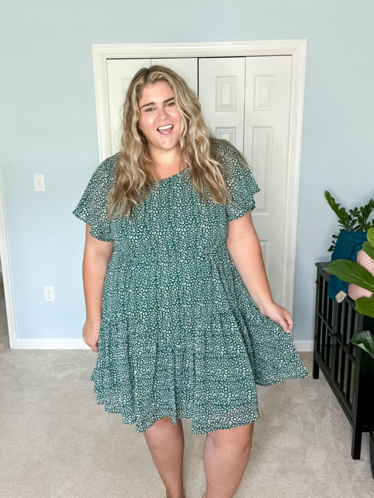 A happy, plus size woman wearing a short, green floral dress; standing in her bedroom. 