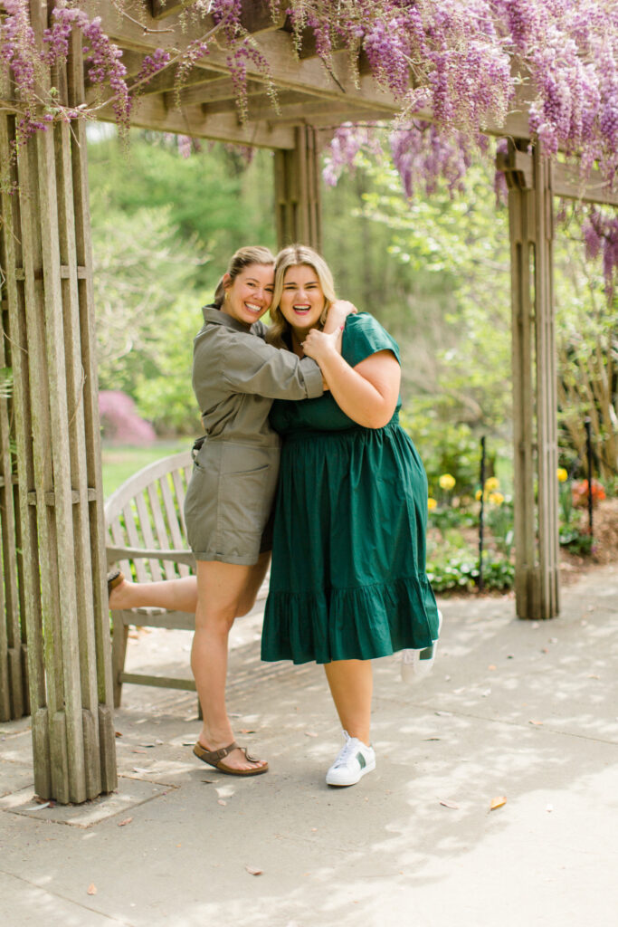 A blonde Caucasian woman in an emerald green dress radiating confidence while being out in nature with her blonde Caucasian sister wearing an army green dress.