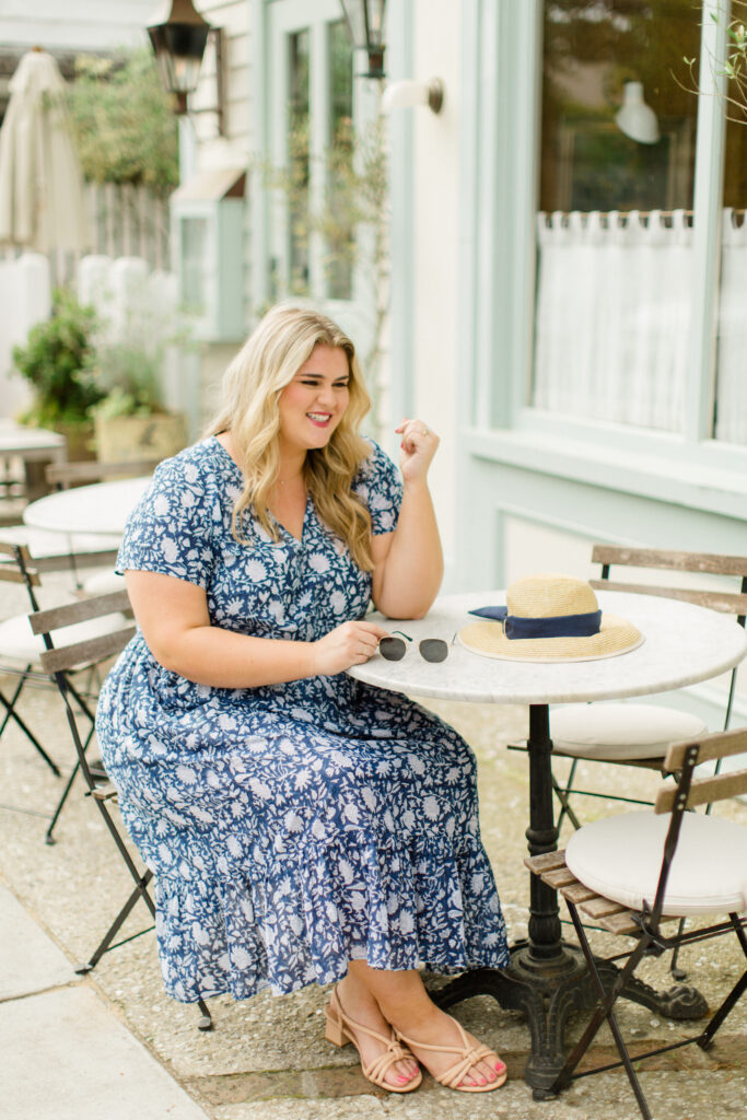 Smiling plus size blonde woman sitting at a café table wearing a blue and white floral dress.